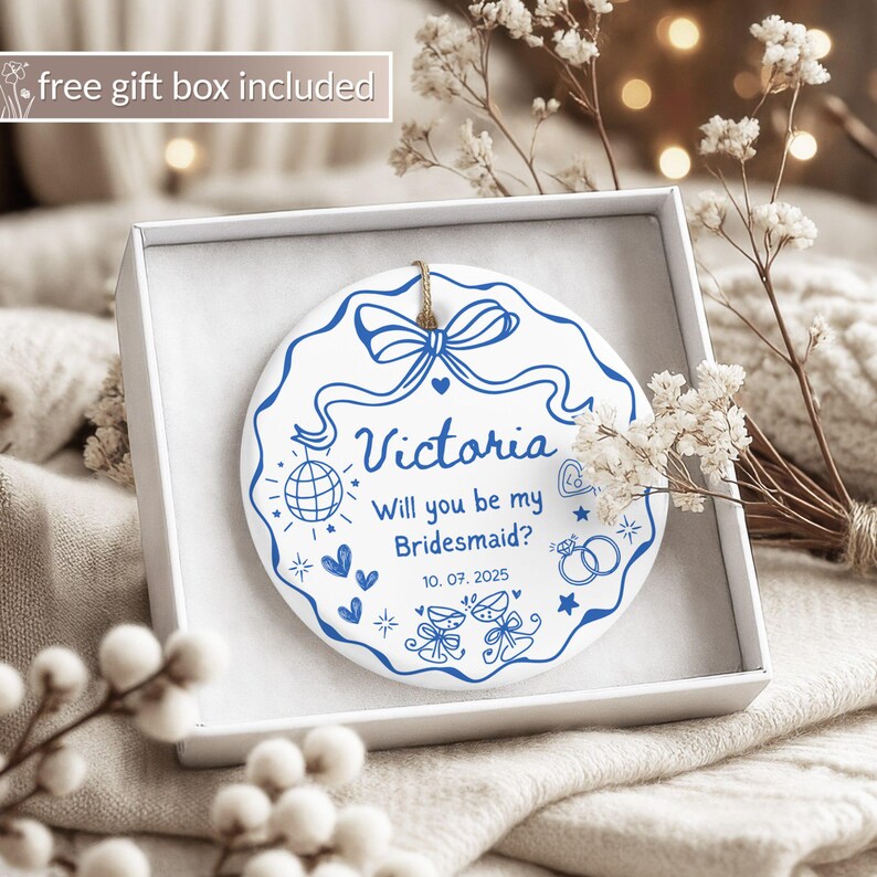 May include: A white ceramic ornament with blue text and designs, presented in a white gift box. The ornament features the name "Victoria" and the question "Will you be my Bridesmaid?" along with a date and decorative elements.