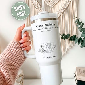 May include: White travel mug with a handle, featuring the text "Cross Stitching because stabbing people is wrong" and a floral and spool design. The mug is held by a person wearing a pink sweater.