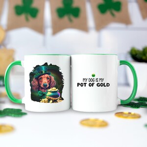 May include: White ceramic mug with a green rim. One side features a cartoon dachshund wearing a green leprechaun hat and holding a pot of gold. The other side says "My dog is my pot of gold".