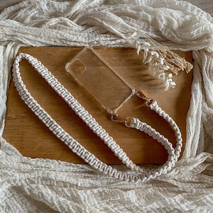 Könnte beinhalten: Eine transparente Handyhülle mit einem Makramee-Handyband in einem natürlichen Off-White-Ton. Der Riemen ist mit goldfarbenen Verschlüssen befestigt. Die Handyhülle und der Riemen liegen auf einer Holzoberfläche mit weißem Stoff und Trockenblumen.