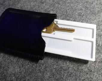 May include: A black leather key holder with a white plastic insert that holds a single key. The key is gold and has a silver keyhole.