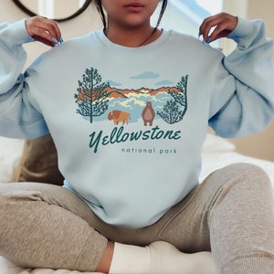 May include: A light blue sweatshirt featuring a graphic design of a bison and a bear in front of a mountain landscape. The text "Yellowstone National Park" is printed below the graphic.
