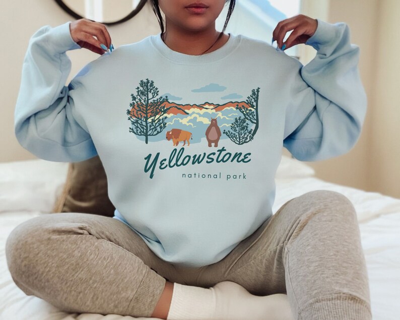 May include: A light blue sweatshirt featuring a graphic design of a bison and a bear in front of a mountain landscape. The text "Yellowstone National Park" is printed below the graphic.