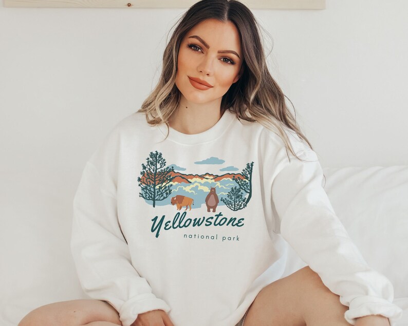 May include: White sweatshirt featuring a graphic design of a mountain landscape, a bison, and a bear. The text "Yellowstone National Park" is printed below the graphic.