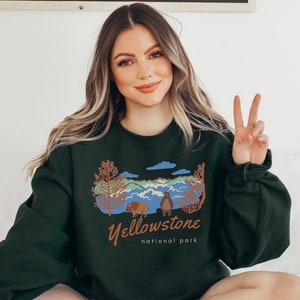 May include: A dark green sweatshirt featuring a graphic print of a mountain landscape, a bear, and a bison. The text "Yellowstone National Park" is printed below the graphic.