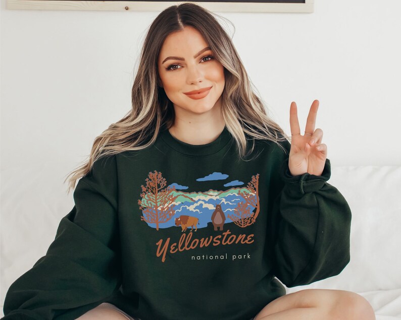 May include: A dark green sweatshirt featuring a graphic print of a mountain landscape, a bear, and a bison. The text "Yellowstone National Park" is printed below the graphic.