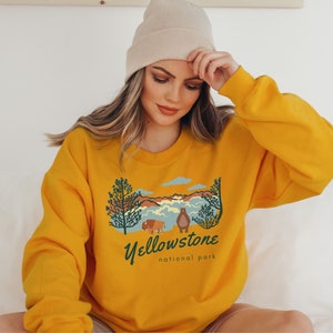 May include: Mustard yellow sweatshirt featuring a vintage-style graphic of a mountain landscape, trees, and a bear and bison. The text "Yellowstone National Park" is printed below the graphic.
