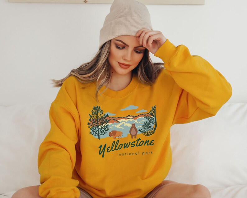 May include: Mustard yellow sweatshirt featuring a vintage-style graphic of a mountain landscape, trees, and a bear and bison. The text "Yellowstone National Park" is printed below the graphic.