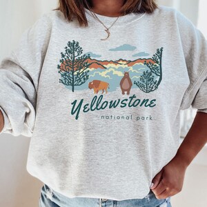May include: A grey sweatshirt featuring a graphic design of a mountain range, a bear, a bison, and trees. The text "Yellowstone National Park" is printed below the graphic.