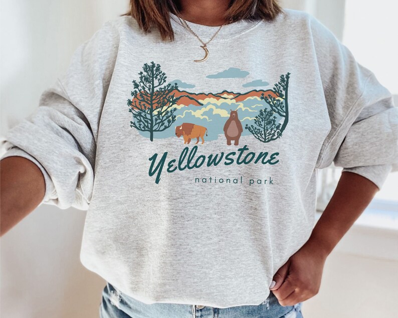 May include: A grey sweatshirt featuring a graphic design of a mountain range, a bear, a bison, and trees. The text "Yellowstone National Park" is printed below the graphic.