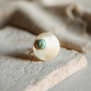 May include: A ring featuring a large, circular, brushed gold-tone metal disc with a smooth, matte finish. A small, round, polished turquoise gemstone with natural brown veining is set off-center on the surface of the gold disc.