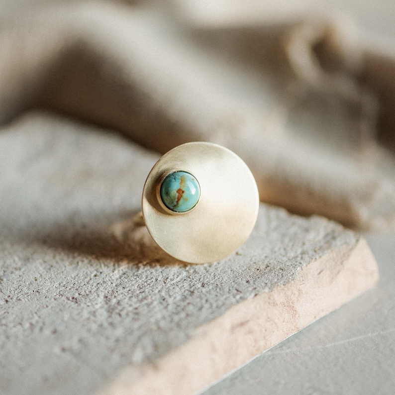 May include: A ring featuring a large, circular, brushed gold-tone metal disc with a smooth, matte finish. A small, round, polished turquoise gemstone with natural brown veining is set off-center on the surface of the gold disc.
