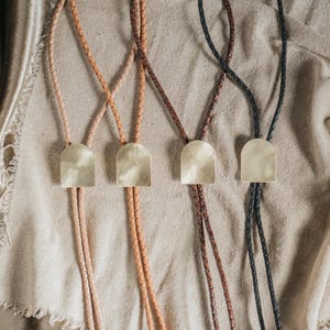 May include: Four bolo ties featuring braided leather cords in shades of tan, light brown, dark brown, and black. Each tie is accented with a polished, arch-shaped metal slide that has a smooth, reflective surface and a rounded top edge.