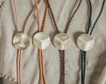 Puede incluir: Cuatro corbatas bolo con cordones de cuero trenzado en tonos beige claro, tostado, marrón oscuro y negro. Cada corbata tiene un pasador circular de metal dorado mate con un diseño de superficie cóncava sutil y tridimensional.