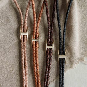 May include: Four braided leather cords in beige, tan, dark brown, and black. Each cord features a sliding gold-toned metal H-shaped clasp that adjusts the length of the loop. The cords are arranged vertically, showcasing the texture of the braided leather.