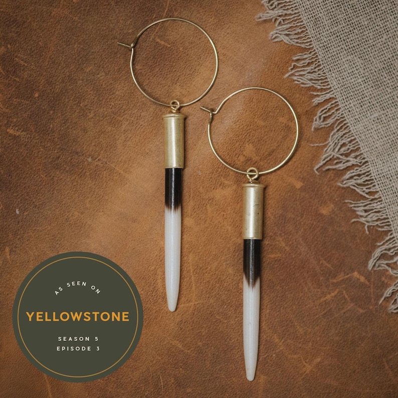 May include: A pair of gold hoop earrings with a long, white and black, bullet-shaped pendant hanging from each hoop. The earrings are on a brown leather surface. The text "As seen on Yellowstone Season 3 Episode 3" is on a green circle.