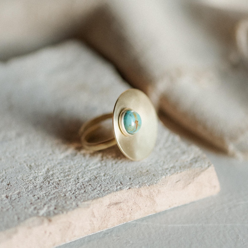 May include: A gold-toned ring featuring a large, flat, circular disc element. A small, round, blue-green turquoise-colored stone is set in the center of the disc. The metal surface has a soft, matte finish, and the band is a simple, thin wire design.
