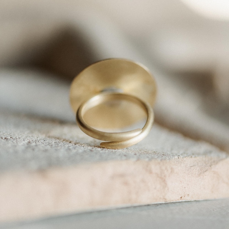 May include: A gold-toned ring featuring a circular flat disc top and an open, adjustable band design. The metal surface has a matte, brushed finish, and the ring is positioned to show the back of the circular element and the overlapping band structure.