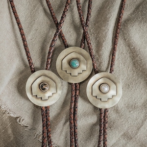 May include: Three bolo ties featuring braided brown leather cords and circular metal slides. Each slide has a central shield-shaped emblem with a gemstone: one with a turquoise stone, one with a dark patterned stone, and one with a white pearl-like bead.