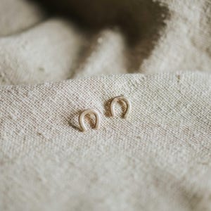 May include: A pair of small, metallic, horseshoe-shaped stud earrings with a smooth, polished finish. The earrings are positioned side-by-side, displaying a simple, curved design that forms an open arch shape, reflecting light on their surface.