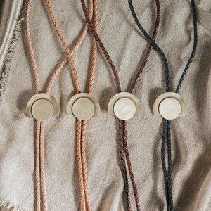 May include: Four bolo ties featuring braided leather cords in shades of tan, light brown, dark brown, and black. Each tie is centered with a circular brass-toned metal slide that features a smooth, flat disc set against a semi-circular backing.