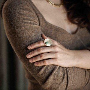 May include: A ring featuring a large, flat, circular gold-toned metal disc with a small, round, turquoise-colored stone set off-center. The metal surface has a smooth, matte finish, and the design is simple, focusing on the contrast between the gold disc and the blue-green stone.
