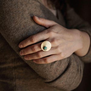 May include: A gold-toned ring featuring a large, flat, circular metal disc as the primary setting. A small, round, blue-green turquoise gemstone is embedded off-center within the smooth, polished surface of the circular gold-colored metal disc.