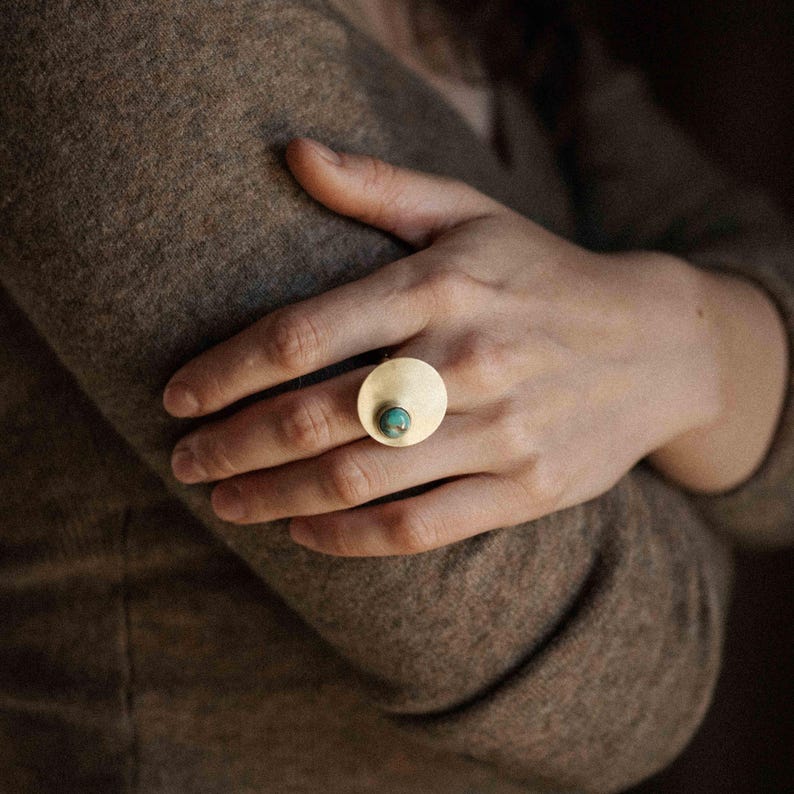 May include: A gold-toned ring featuring a large, flat, circular metal disc as the primary setting. A small, round, blue-green turquoise gemstone is embedded off-center within the smooth, polished surface of the circular gold-colored metal disc.