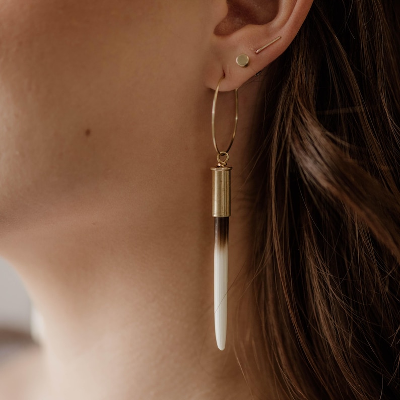May include: Gold hoop earrings with a bullet casing and a long, white and brown bone pendant.