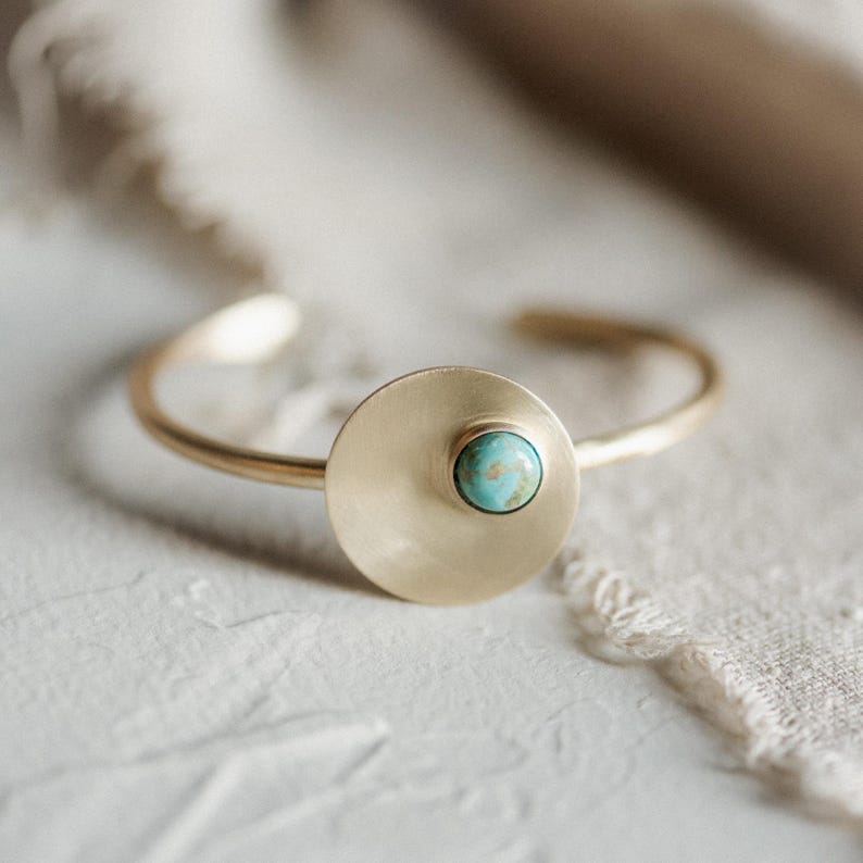 Pu&ograve; includere: Un bracciale a fascia in metallo color oro caratterizzato da un elemento a disco piatto, circolare e liscio. Una piccola pietra rotonda di colore blu-verde turchese &egrave; incastonata decentrata sulla superficie del disco. Il bracciale ha un design aperto.