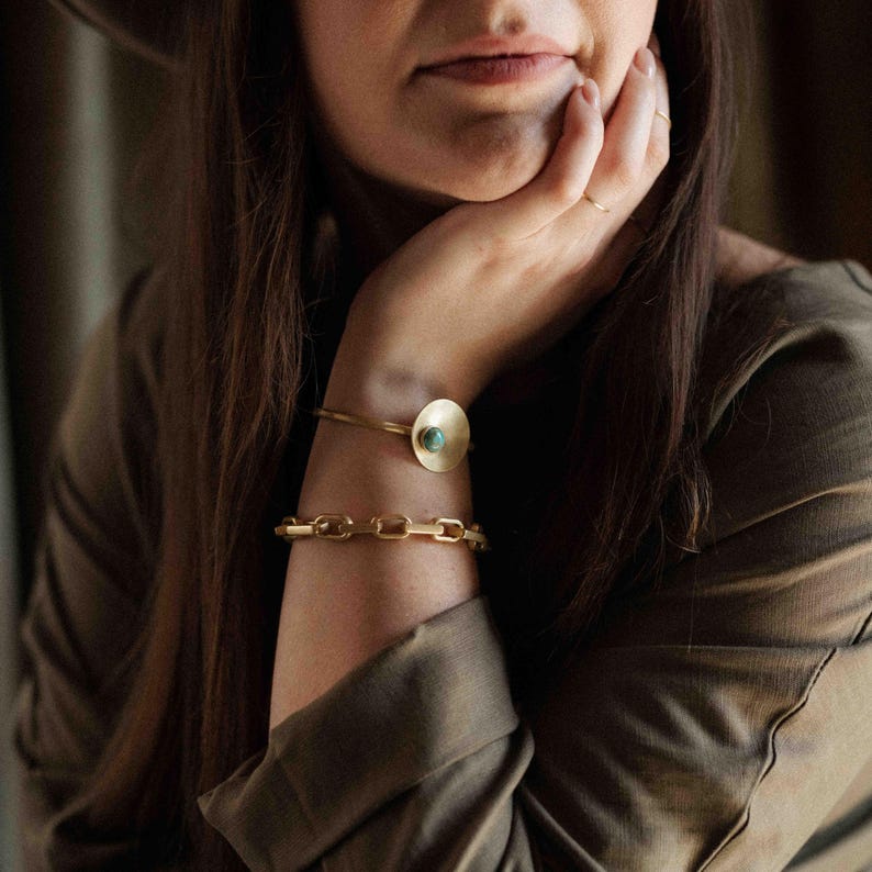 Pu&ograve; includere: Un bracciale rigido dorato con un disco circolare e una piccola pietra incastonata color turchese, abbinato a un bracciale a catena dorato composto da maglie rettangolari, entrambi indossati al polso.