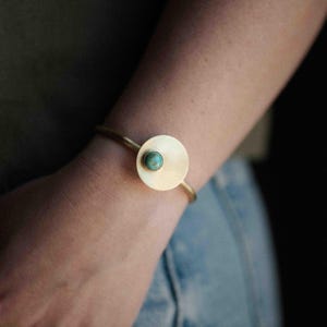 Pu&ograve; includere: Un bracciale a fascia in metallo color oro con un elemento a disco piatto circolare. Il centro del disco &egrave; incastonato con una piccola pietra rotonda di colore blu-verde turchese. Il bracciale ha un design sottile e aperto sul polso.