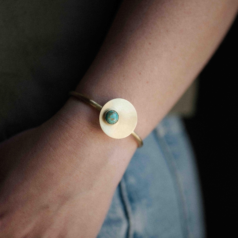 Pu&ograve; includere: Un bracciale a fascia in metallo color oro con un elemento a disco piatto circolare. Il centro del disco &egrave; incastonato con una piccola pietra rotonda di colore blu-verde turchese. Il bracciale ha un design sottile e aperto sul polso.