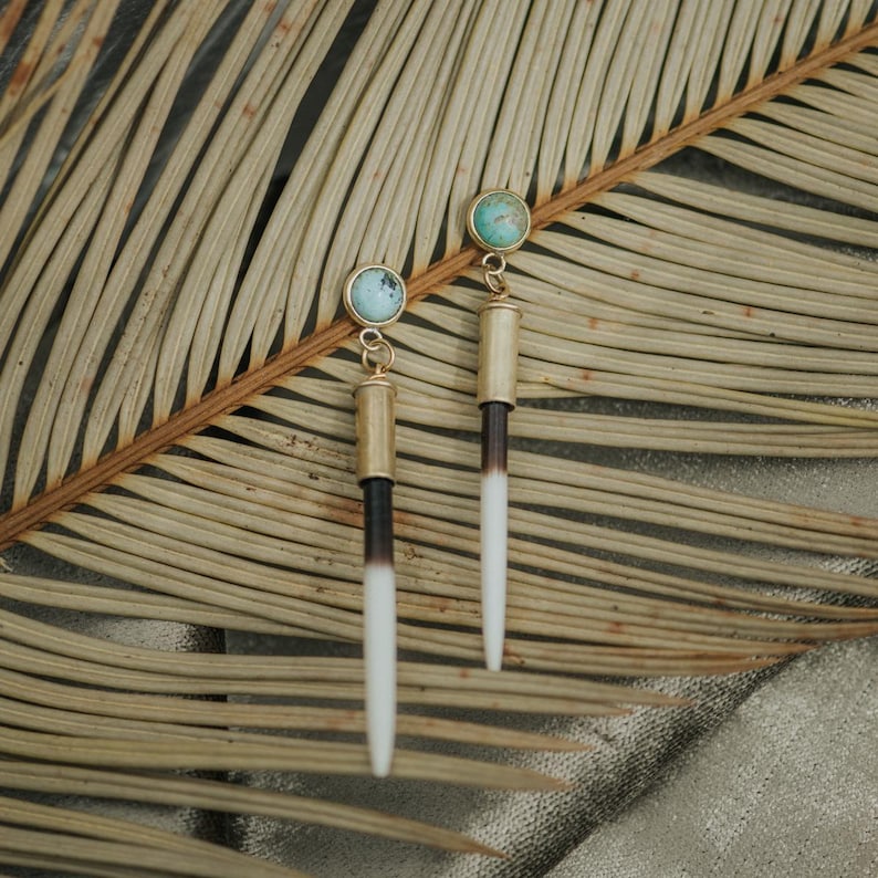 May include: A pair of long, gold and white earrings with turquoise stones. The earrings are hanging from a palm leaf.