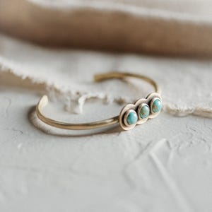 May include: A gold-toned metal cuff bracelet featuring three round, blue-green turquoise stones set in a horizontal row. The stones are encased in individual circular metal settings that are connected to the thin, open-ended band.