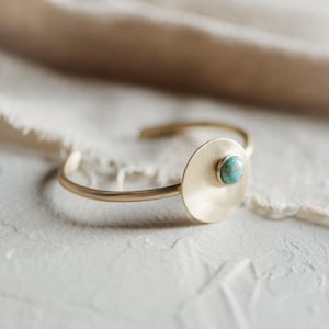 Op de afbeelding: Een goudkleurige metalen armband met een gladde, cirkelvormige platte schijf als accent. In het midden van de schijf zit een kleine, ronde, gepolijste turkoois steen met natuurlijke bruine aders, gezet in een eenvoudige goudkleurige zetting.