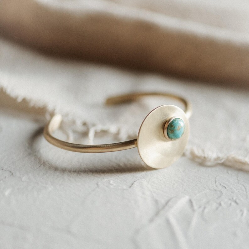 Pu&ograve; includere: Un bracciale rigido in metallo color oro caratterizzato da un disco piatto circolare e liscio. Al centro del disco si trova una piccola pietra turchese rotonda e lucida con venature marroni naturali, incastonata in una semplice montatura dorata.