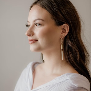 May include: A woman with long brown hair wearing a white shirt and gold hoop earrings with a white and brown pendant.