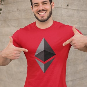 Puede incluir: Un hombre con una camiseta roja con un logotipo gris de la criptomoneda Ethereum impreso en la parte delantera. Está sonriendo y señalando el logotipo con ambas manos.