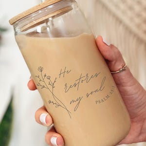 Może przedstawiać: Przezroczysty szklany kubek z drewnianą pokrywką i słomką. Kubek jest wypełniony jasnobrązowym napojem i ma czarno-biały wzór z napisem "He restores my soul Psalm 23:3".