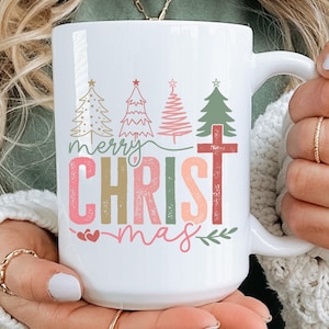 Könnte beinhalten: Weiße Keramik-Tasse mit dem Schriftzug "merry CHRISTmas" in bunten Buchstaben und Weihnachtsbäumen. Die Tasse hat einen Henkel und wird von einer Person gehalten.