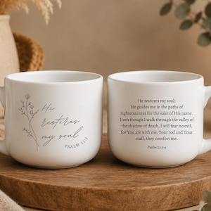May include: Two white ceramic mugs featuring black text and line art. One mug displays the phrase He restores my soul and Psalm 23:3 with a floral sketch. The other mug features the full text of Psalm 23:3-4 in a clear, black serif font.