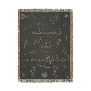 May include: A dark gray woven throw blanket with a floral pattern and the text "Consider the wildflowers" in a light pink color.