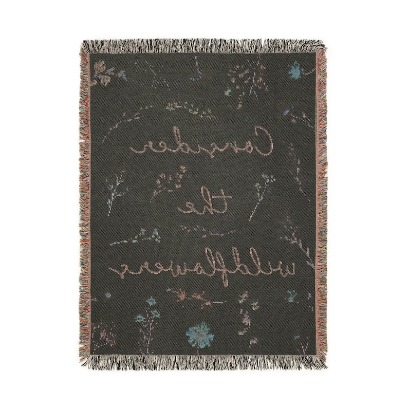 May include: A dark gray woven throw blanket with a floral pattern and the text "Consider the wildflowers" in a light pink color.