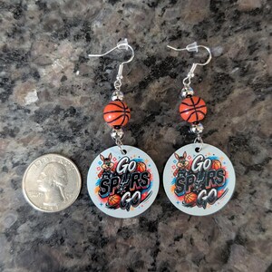 May include: A pair of dangle earrings featuring round white pendants with a graphic of a cartoon coyote, basketballs, and the text Go SPURS Go. Each earring includes a small orange basketball bead and silver-toned hardware, shown next to a US quarter.