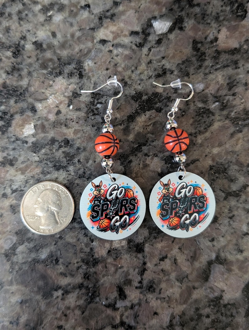 May include: A pair of dangle earrings featuring round white pendants with a graphic of a cartoon coyote, basketballs, and the text Go SPURS Go. Each earring includes a small orange basketball bead and silver-toned hardware, shown next to a US quarter.