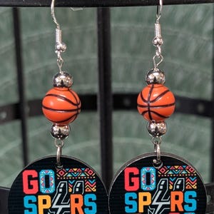May include: A pair of dangle earrings featuring a round black disc with the text GO SPURS GO in blue and orange, accented with a colorful geometric pattern. Above the disc is a silver-toned bead and a small orange basketball-shaped bead.