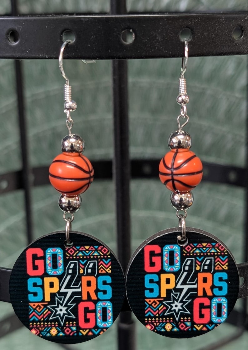 May include: A pair of dangle earrings featuring a round black disc with the text GO SPURS GO in blue and orange, accented with a colorful geometric pattern. Above the disc is a silver-toned bead and a small orange basketball-shaped bead.
