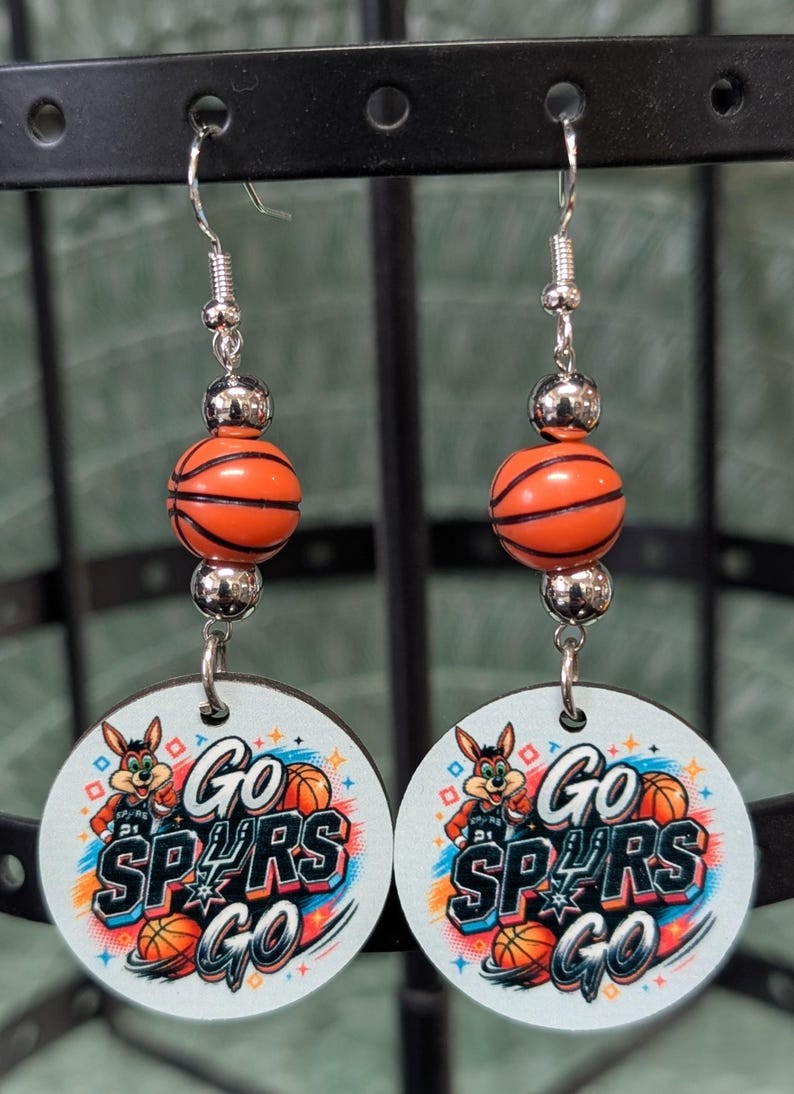 May include: A pair of dangle earrings featuring a round white disc with a cartoon mascot, basketballs, and the text "GO SPURS GO" in black and blue. Each earring includes a small orange basketball bead flanked by two silver-toned metallic beads.