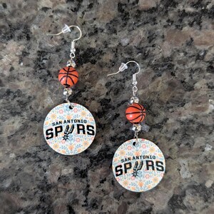 May include: A pair of dangle earrings featuring silver-toned hooks, small orange basketball beads, and circular charms. The charms display a floral pattern in soft colors with the text SAN ANTONIO SPURS printed in black, including the team logo.